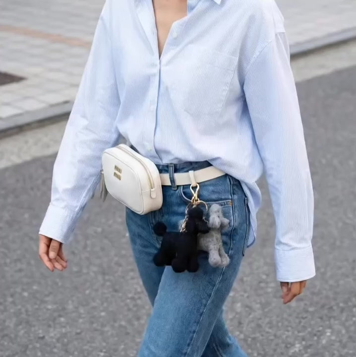 Four handmade needle felted wool poodle keychains in white, brown, grey, and black colors displayed on cream fabric with wooden toys