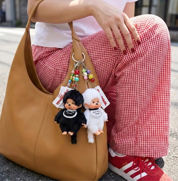 Black and white Monchhichi plush keychains layered with colorful bead keychains on a camel leather tote bag, model wearing red check trousers