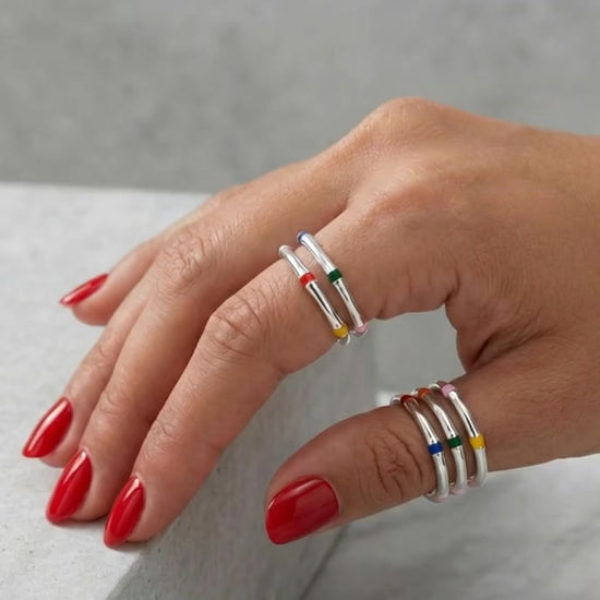 Model with red nail polish wearing layered multicolor enamel 925 silver stacking rings on two fingers, marble background