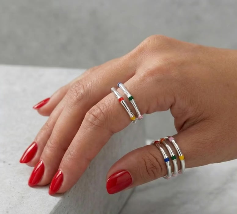 Model with red nail polish wearing layered multicolor enamel 925 silver stacking rings on two fingers, marble background