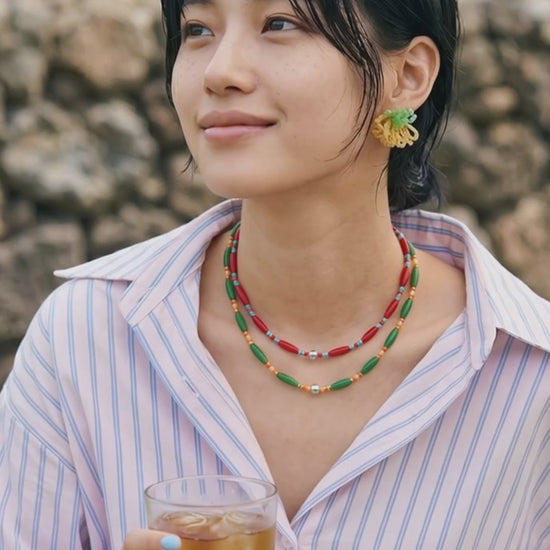 A stylish young woman holding a glass of iced tea outdoors. She wears an oversized pink and blue striped shirt styled with mint green beaded cluster earrings, epitomizing chic Korean casual wear.