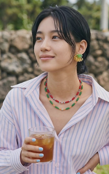 A stylish young woman holding a glass of iced tea outdoors. She wears an oversized pink and blue striped shirt styled with mint green beaded cluster earrings, epitomizing chic Korean casual wear.